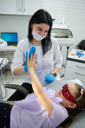 Foto de Sonriente dentista doctora confiada y niña pequeña paciente con gafas UV, dar cinco altos, sentado en la silla de dentistas, feliz después del tratamiento de los dientes y regulat chequeo dental en la clínica de odontología - Imagen libre de derechos