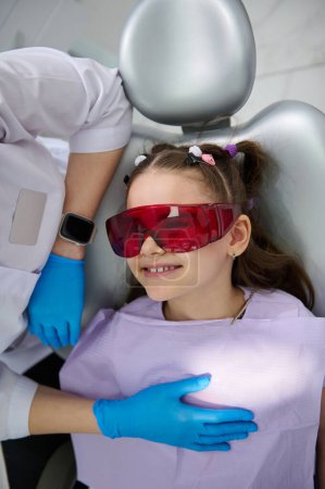 Téléchargez les photos : Gros plan sur le dessus d'une petite fille heureuse et souriante, patiente portant des lunettes UV, dentiste visiteuse à la clinique de dentisterie pédiatrique. Santé dentaire et hygiène buccodentaire. Le concept de traitement des dents de bébé - en image libre de droit