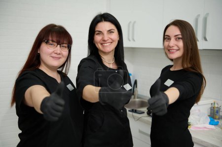 Téléchargez les photos : Équipe confiante de femmes dentistes médecins orthodontistes en uniforme médical noir, souriant largement regarder la caméra, geste avec les pouces levés, montrant signe des mains d'approbation, dans un intérieur de bureau dentaire - en image libre de droit