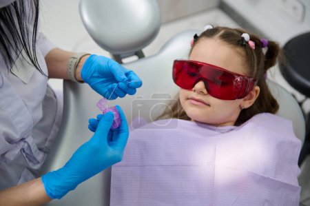 Téléchargez les photos : Enfant fille mignonne écoute un hygiéniste dentaire expliquant comment nettoyer les dents à l'aide d'un fil dentaire après le brossage des dents, lors d'un rendez-vous chez le dentiste à la clinique dentaire. Concept de soins buccodentaires et d'hygiène - en image libre de droit
