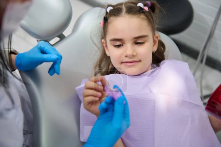 Téléchargez les photos : Portrait en gros plan d'une belle petite fille du Moyen-Orient à un rendez-vous chez le dentiste, tenant un fil dentaire, faisant examiner ses dents dans une clinique de dentisterie pédiatrique. Concept de soins dentaires et d'hygiène - en image libre de droit