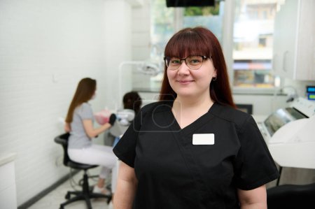 Téléchargez les photos : Portrait confiant d'une jeune dentiste caucasienne heureuse et positive, orthodontiste en uniforme médical noir, souriante regardant la caméra, debout dans le cabinet dentaire d'une clinique dentaire moderne - en image libre de droit