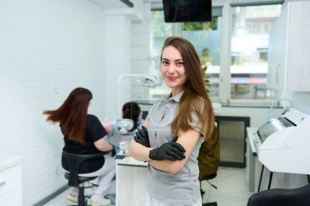 Téléchargez les photos : Femme de charme caucasienne de 25 à 20 ans, portant un uniforme médical gris, médecin dentiste confiante, orthodontiste, chirurgien maxillofacial sourit à la caméra, se tient avec les bras croisés dans une clinique dentaire - en image libre de droit