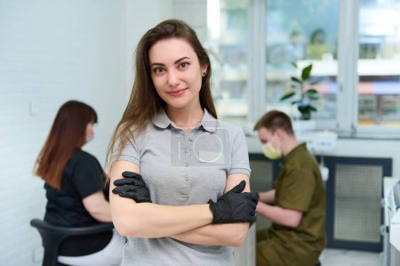 Téléchargez les photos : Portrait confiant d'une jeune dentiste caucasienne engagée avec succès regardant la caméra, debout dans un cabinet dentaire, sur fond d'hygiéniste dentaire et d'assistante travaillant avec un patient - en image libre de droit