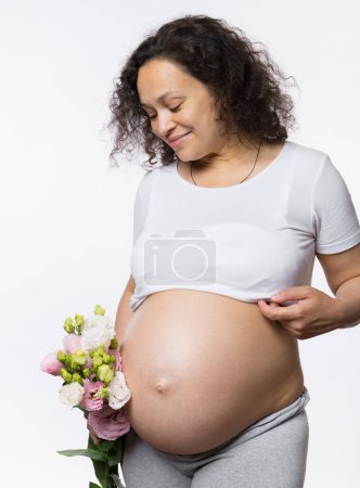 Téléchargez les photos : Belle femme gravide heureuse multi ethnique, future mère adulte tenant un bouquet de fleurs de printemps, souriant en regardant son ventre enceinte à la fin de la grossesse, isolée sur fond de studio blanc - en image libre de droit