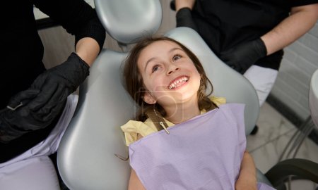 Foto de Retrato de cerca de una adorable niña pequeña sentada en la silla del dentista, sonriendo con una hermosa sonrisa dentada después del chequeo dental en la clínica de odontología. Cuidado bucal e higiene dental - Imagen libre de derechos