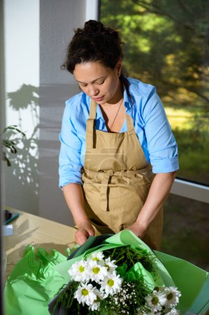 Photo for Curly haired charming multi-ethnic adult pregnant woman florist, in beige work apron, enjoying her creative hobby in pregnancy time, arranging flower bouquets in floral design studio. Small business - Royalty Free Image