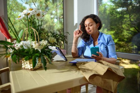 Photo for Multi-ethnic confident thoughtful perplexed woman florist, event manager, businesswoman, entrepreneur holding mobile phone, working in her workshop in a floral design studio. Small business. Startup - Royalty Free Image