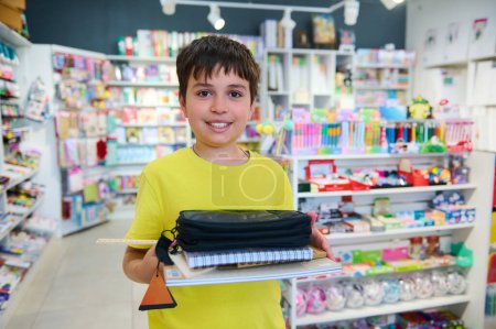 Téléchargez les photos : Adorable écolier diligent recueillant des fournitures scolaires dans un magasin de papeterie, souriant en regardant la caméra. Retour à l'école. Préparation de la nouvelle année académique - en image libre de droit