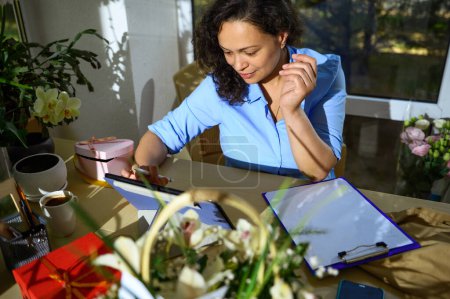 Photo for Happy Latin American woman using tablet, works online, receives orders, sitting at desk with clipboard, surrounded by exotic flowers and fresh plants, in a floral design studio. Arrangements and gifts - Royalty Free Image