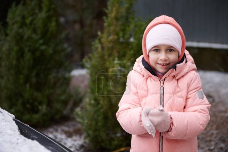 Photo for Mischievous little child girl with rosy cheeks, wearing pink warm down jacket, cutely smiles looking at camera, makes snowballs while playing in a snowy backyard. Winter holidays. Nordic lifestyle - Royalty Free Image
