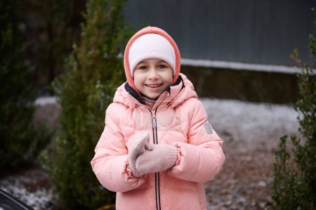 Photo for Adorable preschooler little child girl with rosy cheeks, wearing pink warm down jacket, cutely smiles looking at camera, makes snowballs while playing in a snowy backyard. Wintertime. Nordic lifestyle - Royalty Free Image