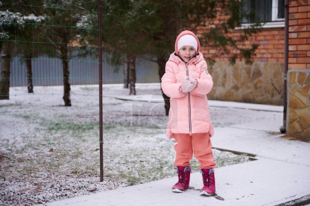 Photo for Full-length portrait of adorable little kid girl with rosy cheeks, wearing warm pink winter clothes, playing in a snowy backyard, smiling, looking at camera. Winter and christmas holidays concept - Royalty Free Image