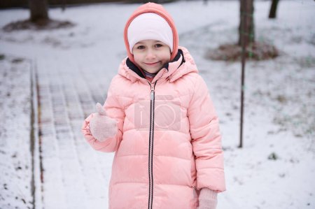 Photo for Caucasian lovely little kid girl with rosy cheeks, wearing pink warm down jacket, smiles looking at camera, gestures with thumb up while walking in a snow covered nature. Wintertime. Nordic lifestyle - Royalty Free Image