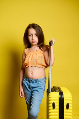 Téléchargez les photos : Belle petite fille caucasienne de 6 ans, voyageuse portant un haut orange vif et un jean bleu, regardant avec confiance la caméra, posant avec une valise jaune, isolée sur fond jaune - en image libre de droit