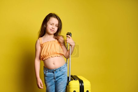 Photo for Cute Caucasian girl traveling abroad, going for weekend getaway, posing with yellow suitcase over yellow background, winking at camera. Children. Travel. Journey. Tourism. Summer. Holidays. Vacation - Royalty Free Image
