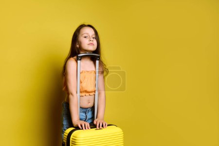 Téléchargez les photos : Fille enfant heureux dans des vêtements d'été lumineux élégants, avec valise jaune, regardant la caméra, isolé sur fond jaune. Enfants voyageant seuls pour étudier à l'étranger ou des vacances d'été. Voyage. Vol aérien - en image libre de droit