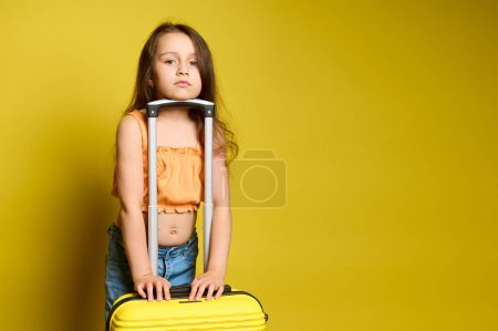 Foto de Hermosa niña caucásica en elegante top naranja de verano y pantalones vaqueros azules casuales, de pie cerca de su vibrante maleta amarilla de moda, mirando a la cámara, aislado en el fondo del estudio amarillo - Imagen libre de derechos