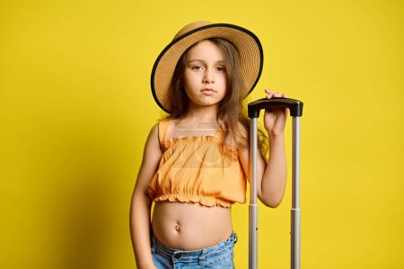 Photo for Serious little kid girl in trendy summer wear, with suitcase, travelling abroad for weekend getaway, isolated on bright yellow studio background. Travel and booking agency. Trip. Journey. Air flight - Royalty Free Image
