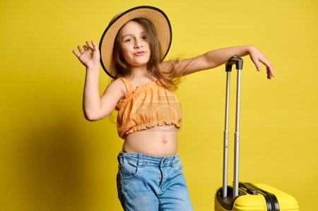 Téléchargez les photos : Mischievous gaie petite fille à la mode avec valise à la mode, portant un chapeau de paille d'été, haut orange jean bleu nad, voyageant à l'étranger pour les vacances d'été. Voyage. Voyage. Voyage. Vol aérien - en image libre de droit