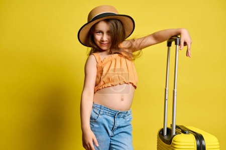 Téléchargez les photos : Joyeux enfant fille sourit joliment à la caméra, posant avec une élégante valise jaune, vêtue d'un haut orange vif, un jean bleu et un chapeau de paille, isolé sur fond jaune. Voyage, enfants et concept d'été - en image libre de droit