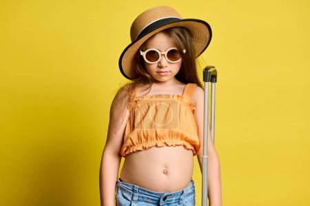 Photo for Caucasian adorable child girl, little tourist passenger in stylish summer wear, straw hat and sunglasses, posing with suitcase, looking confidently at camera, isolated on yellow background. Tourism - Royalty Free Image