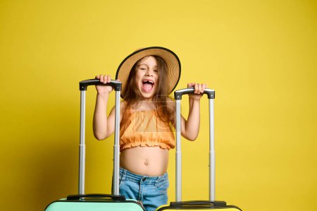 Téléchargez les photos : Petite fille drôle caucasienne espiègle avec des valises, portant un chapeau de paille d'été, un haut orange et un jean bleu, souriant en regardant la caméra, profite de son prochain vol, isolée sur fond jaune - en image libre de droit