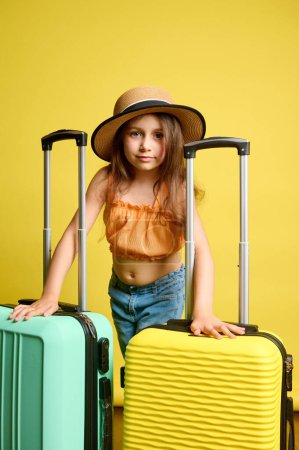 Téléchargez les photos : Enfant à la mode, écolière de 6 ans en chapeau de paille, avec des valises vert clair et jaune vibrantes, voyageant à l'étranger, allant pour une escapade de week-end, regardant avec confiance la caméra, arrière-plan isolé - en image libre de droit