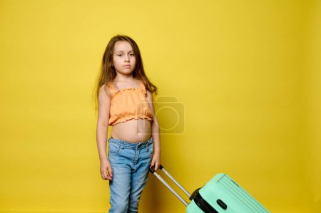 Téléchargez les photos : Adorable petite fille blanche 6 ans, en vêtements d'été tendance, avec une valise élégante vert clair, isolée sur fond de studio jaune. Le concept de voyage, voyage, voyage, week-end, vol aérien - en image libre de droit
