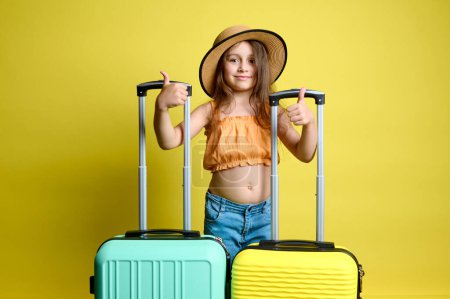 Smiling little tourist traveler passenger in stylish casual summer clothes, gestures with thumbs up, standing with yellow and light green polycarbonate, plastic suitcases, isolated on studio backdrop