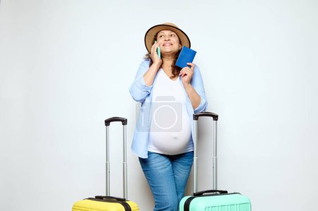 Happy smiling pregnant woman, expectant mother talking on smartphone, standing with plastic suitcases and boarding pass, awaiting her flight, expressing positive emotions, isolated on white background