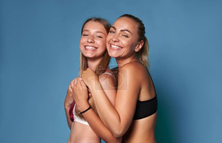 Two Caucasian young women, mom and daughter in underwear, smiling with beautiful toothy smile, hugging each other, isolated studio background. Dental health. Oral hygiene. Teeth bleaching Whitening