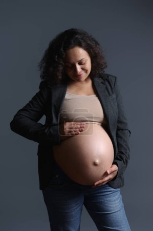 Stylish elegant multi-ethnic, curly haired pregnant woman smiling while gently caressing her big belly in pregnancy 36 week, feeling happy emotions from baby kicks, isolated on gray studio background