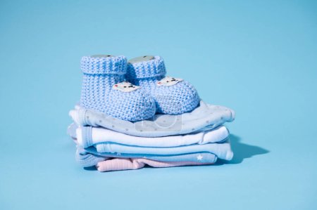 Photo for Still life blue knitted baby boots on a pile of clean laundered ironed newborn bodysuits, isolated on blue pastel background. Preparations of the bag for maternity hospital and child birth. Pregnancy - Royalty Free Image