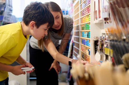 Téléchargez les photos : Adorable écolier adolescent aux cheveux bruns demandant conseil à un directeur des ventes dans un magasin d'art, choisissant des stylos feutre de l'étagère avec un large assortiment d'outils de dessin, de peinture et de croquis colorés - en image libre de droit