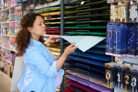 Photo for Multi-ethnic curly-haired pregnant woman in blue casual shirt, taking out a color design paper sheet from the shelf, shopping in an art store or school stationery shop. People. Hobbies and leisures. - Royalty Free Image