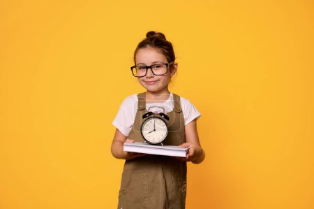 Photo for Authentic smiling little kid girl in eyeglases and stylish casual dress, looking at camera carrying a textbook and alarm clock, isolated on orange studio background. Copy ad space. People. Education - Royalty Free Image