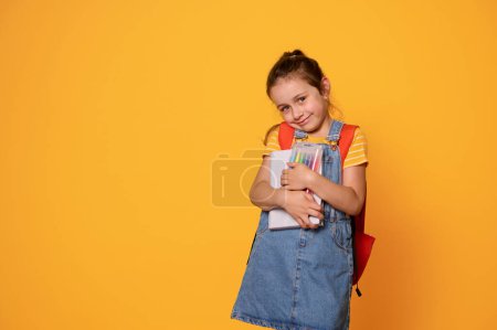 Téléchargez les photos : Caucasienne âge élémentaire mignonne petite fille, première année, élève du primaire portant son sac à dos, étreint ses fournitures scolaires et sourit mignonne, regardant la caméra, fond isolé studio jaune - en image libre de droit
