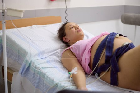 Pregnant woman lying on the bed in comfortable ward, having contractions, checking the heartbeat and pulse of her baby, using an electrocardiogram belt around her belly, in modern maternity hospital.