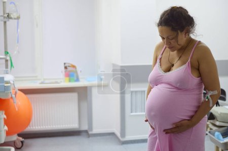 Foto de Mujer embarazada multiétnica de mediana edad, madre biológica con contracción dolorosa, en la sala del hospital, tocando el vientre y mirando hacia abajo. Proceso de parto en el hospital de maternidad. Embarazo 40 semanas - Imagen libre de derechos