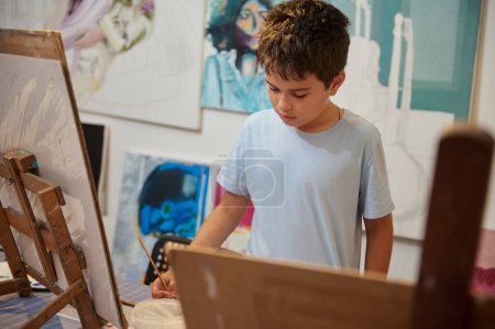 Foto de Niño adolescente confiado sosteniendo pincel, mezclando pinturas, dibujo sobre lienzo en un estudio de arte creativo. Entretenimiento y educación para niños. Artes visuales, Desarrollo de la creatividad, habilidades artísticas e imaginación - Imagen libre de derechos