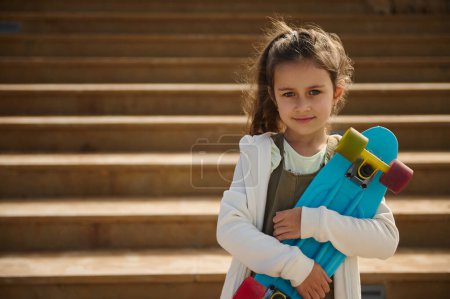 Téléchargez les photos : Authentique portrait de charmante jeune fille caucasienne de 6 ans, tenant skateboard, sourit mignon en regardant la caméra, debout contre fond de marches en marbre beige, vêtu de capuche blanche et salopettes - en image libre de droit