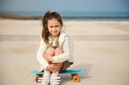 Téléchargez les photos : Portrait grandeur nature d'une petite fille en tenue décontractée, assise sur son penny board sur une aire de jeux sur la promenade maritime sur fond d'océan, souriant en regardant la caméra. Les gens. Les enfants. Mode de vie - en image libre de droit