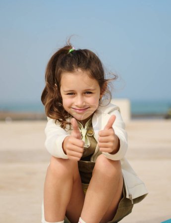 Téléchargez les photos : Portrait d'une authentique petite fille charmante montrant les pouces levés et souriant en regardant la caméra, exprimant l'accord et les émotions positives, assise sur son skateboard à l'extérieur sur fond de mer - en image libre de droit