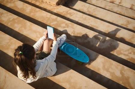 Téléchargez les photos : Vue de dessus d'une jolie petite fille à l'aide d'un smartphone avec écran numérique blanc, assis à l'extérieur et mettant ses pieds sur une planche à roulettes bleue. Les gens. Mode de vie. Communication - en image libre de droit