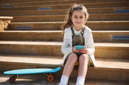 Téléchargez les photos : Authentique portrait de style de vie d'une charmante petite fille, élève de l'école primaire en tenue décontractée, tenant un téléphone portable, assise sur les marches avec son skateboard, sourit en regardant avec confiance la caméra - en image libre de droit