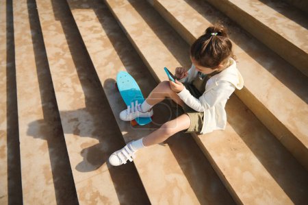 Téléchargez les photos : Vue aérienne d'une enfant fille à l'aide d'un smartphone moderne assis sur des marches à l'extérieur, mettre le pied sur le skateboard, profiter d'un week-end actif heureux à l'extérieur par une chaude journée ensoleillée. Les enfants. Communication en ligne - en image libre de droit