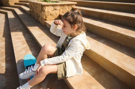 Téléchargez les photos : Adorable enfant de maternelle caucasienne regardant au loin, assise à l'extérieur sur l'étape, se reposant après le skateboard sur une aire de jeux, vêtue d'un sweat à capuche beige et d'une robe kaki - en image libre de droit