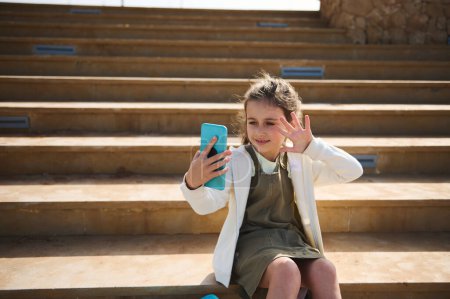 Téléchargez les photos : Portrait confiant d'une enfant heureuse en vêtements décontractés, parlant via un lien vidéo, faisant du selfie, assise sur des marches à l'extérieur par une chaude journée ensoleillée. Les gens. Communication en ligne sur Internet. Technologie numérique - en image libre de droit