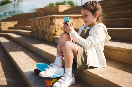 Téléchargez les photos : Portrait style de vie d'une adorable petite fille heureuse avec planche à roulettes, assise sur des marches à l'extérieur, utilisant un smartphone, vérifiant le contenu des médias sociaux, regardant des dessins animés, jouant à un jeu vidéo en ligne - en image libre de droit